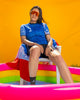 A woman wearing a blue cycling jersey with a geometric wave-inspired design, sitting on a colorful structure against an orange wall.