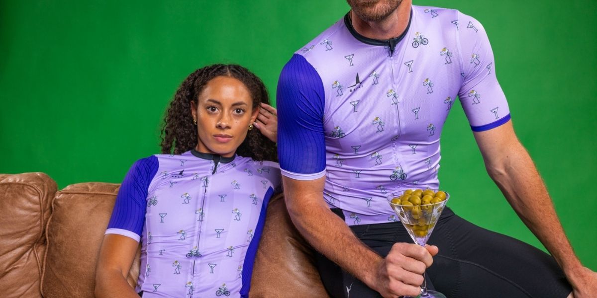 Two people wearing matching purple and blue cycling outfits sitting on a couch with a green screen background.