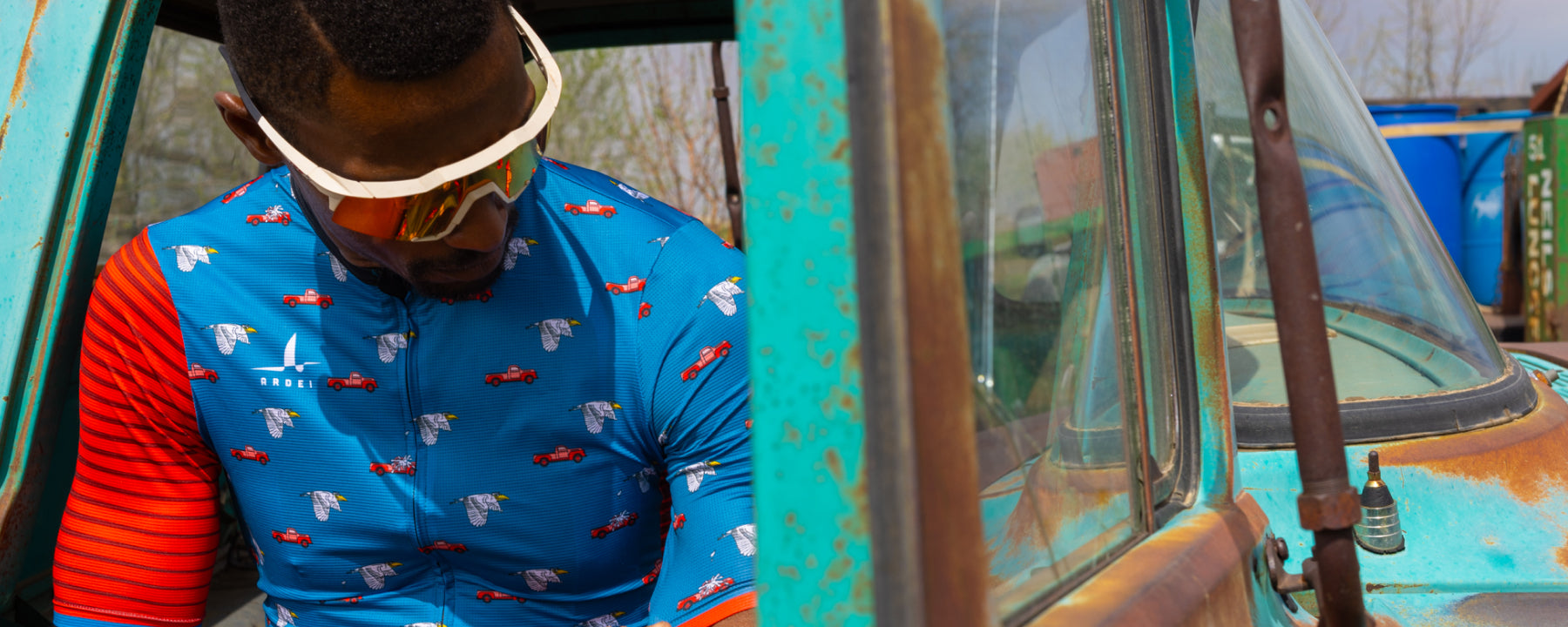 Male cyclist wearing Ardei blue and red cycling jersey with car and wing graphics, leaning on a retro truck backdrop 