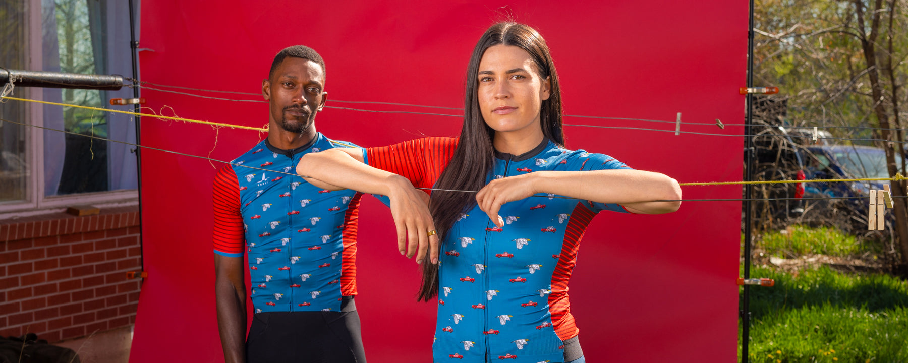 Male and female cyclists wearing matching Ardei blue and red cycling jerseys with car and wing graphics, standing in front of a red backdrop with clotheslines in backyard setting