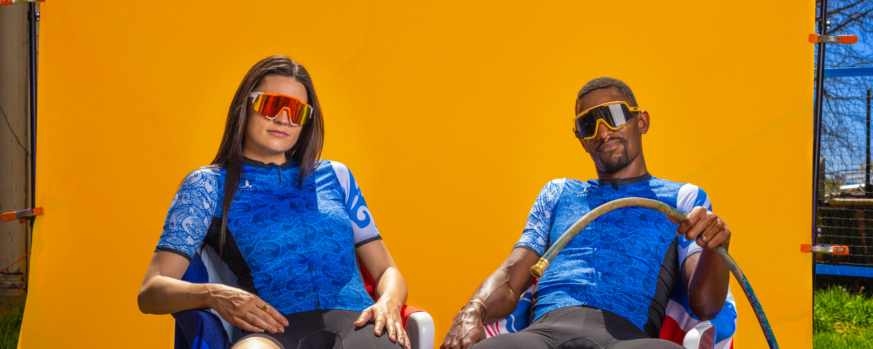 Two cyclists wearing matching Ardei blue summer cycling jerseys, sitting outdoors in front of a bright orange backdrop, wearing performance sunglasses