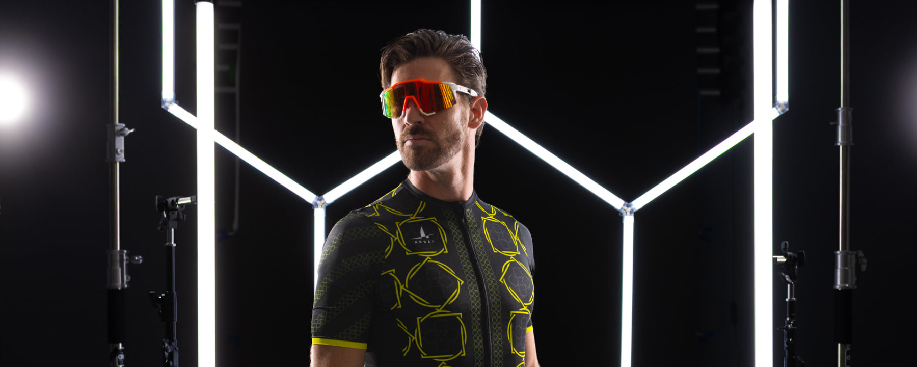 Male cyclist wearing Ardei summer cycling jersey with neon yellow pattern and performance sunglasses, studio lighting setup in background