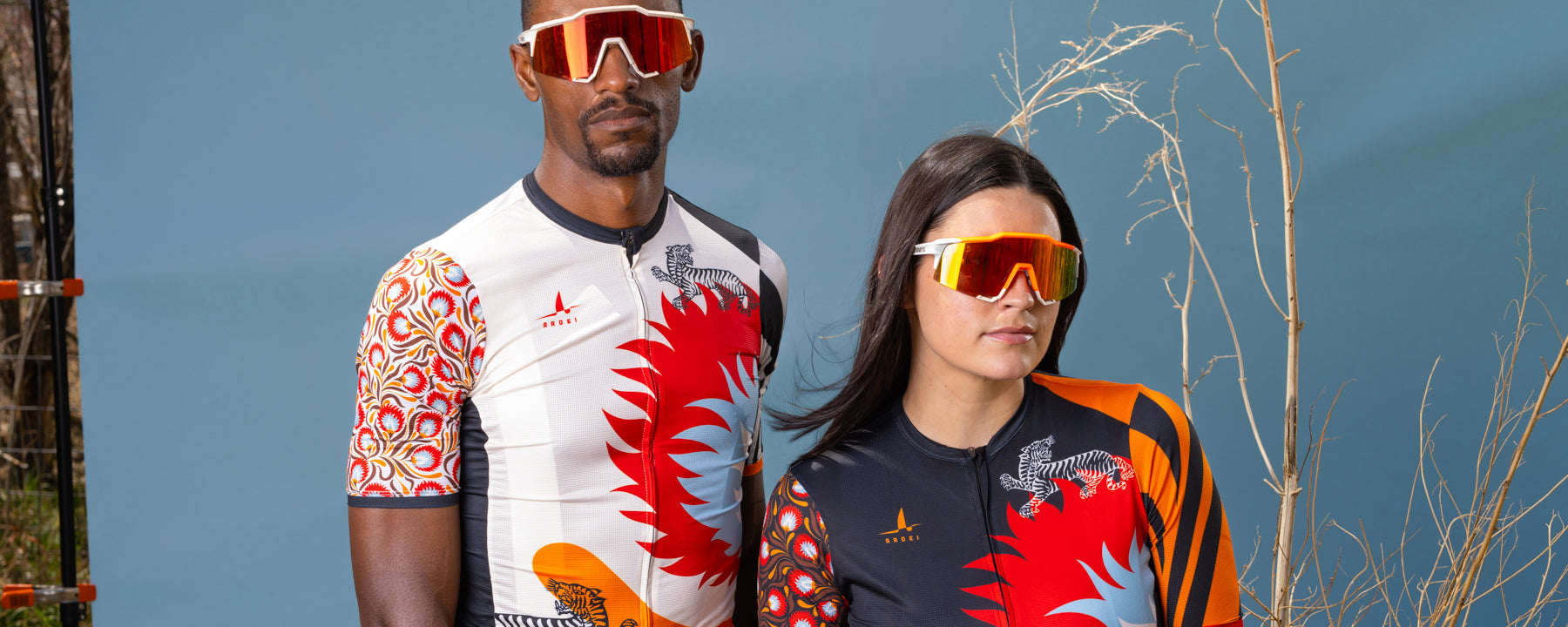 Male and female cyclists wearing Ardei white floral and black floral cycling jerseys with fierce tiger graphics in front of a blue backdrop and a savannah bush